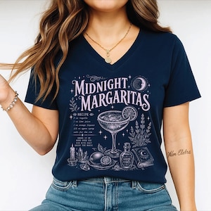 May include: A navy blue V-neck t-shirt featuring a light purple graphic print with the text Midnight Margaritas. The design includes a margarita glass, a recipe list, a candle, crystals, a small bottle labeled Good Vibes, and a book.