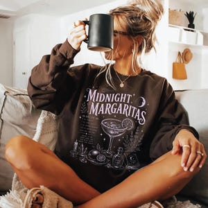May include: A dark brown long-sleeve sweatshirt featuring a white graphic print that reads Midnight Margaritas, accompanied by illustrations of a cocktail glass, lime slices, a small bottle, a candle, crystals, and a book with a moon symbol.
