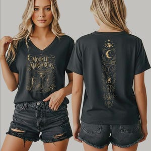 May include: A dark gray V-neck t-shirt featuring a gold graphic on the front with the text "MOONLIT MARGARITAS" above a margarita glass, coffee pot, and citrus slices. The back displays a vertical gold design of moons, a glass, and a key.