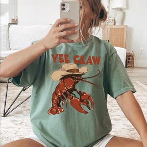 A woman is sitting on the floor, taking a selfie with her phone, wearing a green t-shirt with a lobster graphic and the text "YEE CLAW" printed on it.