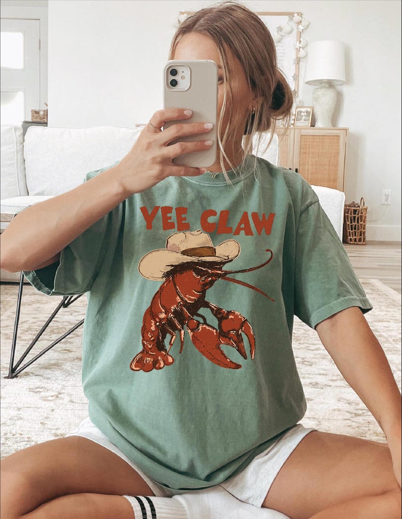 A woman is sitting on the floor, taking a selfie with her phone, wearing a green t-shirt with a lobster graphic and the text "YEE CLAW" printed on it.