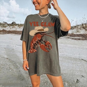 A woman wearing a gray t-shirt with a graphic of a lobster and the text "YEE CLAW" is standing on a beach.