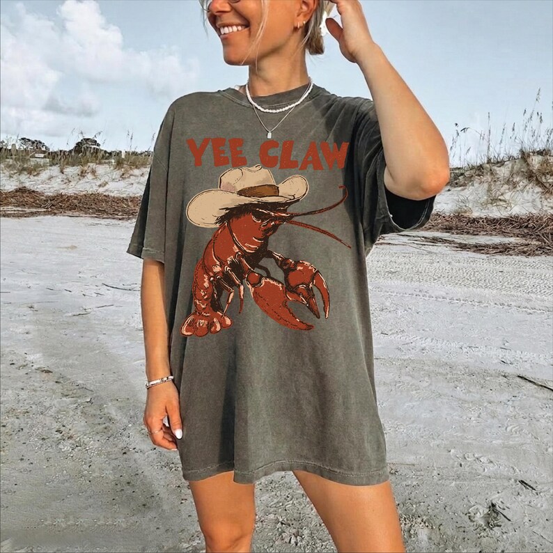 A woman wearing a gray t-shirt with a graphic of a lobster and the text "YEE CLAW" is standing on a beach.