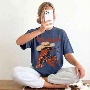 A woman is sitting cross-legged on the floor, holding a smartphone up to her face, with a t-shirt featuring a lobster graphic and the text "YEE CLAW" visible.