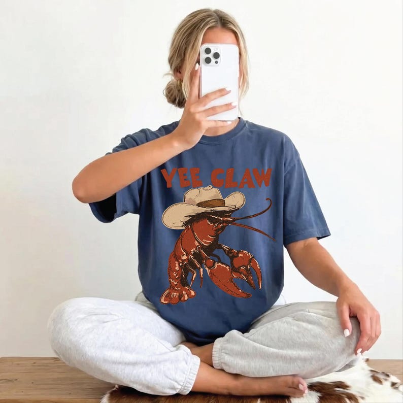 A woman is sitting cross-legged on the floor, holding a smartphone up to her face, with a t-shirt featuring a lobster graphic and the text "YEE CLAW" visible.