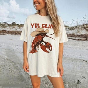 A woman wearing a white t-shirt with a graphic of a lobster and the text "YEE CLAV" printed on it, standing on a beach.