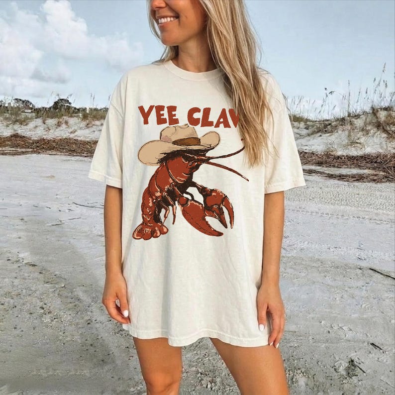 A woman wearing a white t-shirt with a graphic of a lobster and the text "YEE CLAV" printed on it, standing on a beach.