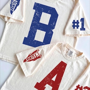 May include: Two off-white t-shirts with distressed graphics. The larger shirt features a blue letter B, a blue pennant with the text MASCOT, and blue #1 text. The smaller shirt features a red letter A, a red pennant with the text MASCOT, and red #3 text.