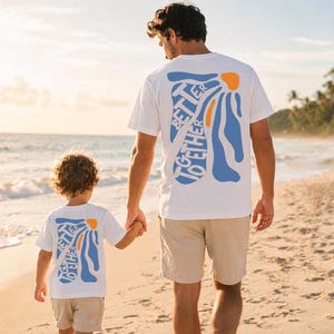 May include: White t-shirts with a blue and orange graphic that reads "Better Together." A man and child are walking on a sandy beach, holding hands. The sun is setting in the background, casting a warm glow over the scene.
