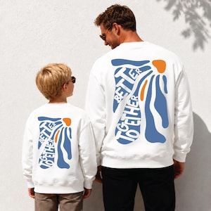 May include: White sweatshirts with a blue and orange graphic that reads "TOGETHER BETTER" on the back. The sweatshirts are worn by a person and a child. The design features abstract shapes and text, creating a matching set.
