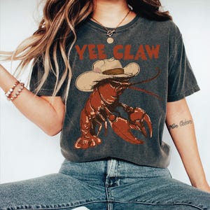 A woman wearing a gray t-shirt with a lobster graphic and the text "YEE CLAW" printed on it.