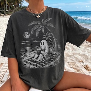 May include: A charcoal gray short-sleeve t-shirt featuring a white graphic print of a ghost sitting on a beach towel under a palm tree, drinking from a coconut. The scene includes a full moon, stars, and ocean waves, rendered in a detailed, sketch-like style.