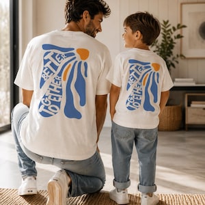 May include: Two white t-shirts with the text "BETTER TOGETHER" in blue and orange, worn by a man and a child. The man is kneeling, and the child is standing, both wearing blue jeans and white sneakers. The shirts have a floral design.