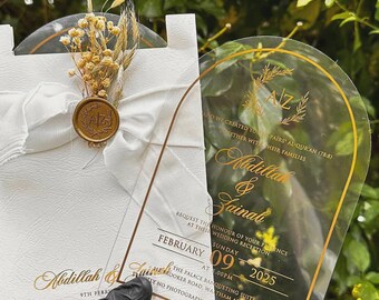 Pode incluir: Convite de casamento com um cartão de acrílico transparente e um cartão branco. O cartão de acrílico tem letras douradas com os nomes Abdillah & Zainab, a data e os detalhes da recepção. O cartão branco tem uma fita e flores secas.