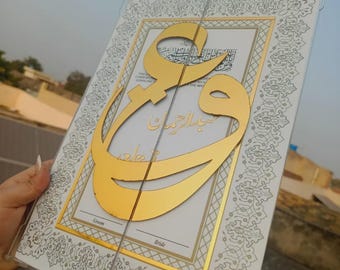 May include: A clear acrylic wedding keepsake with gold accents. The design features gold Arabic calligraphy over a white background with decorative borders. The words "Groom" and "Bride" are visible at the bottom.