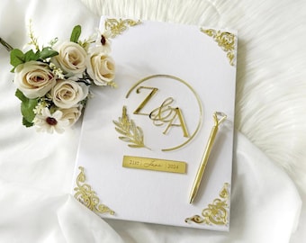 May include: A white wedding guest book with gold accents, including corner designs, initials, and a date. A gold pen with a diamond-shaped top rests beside the book. A bouquet of cream-colored roses and greenery is in the upper left corner.