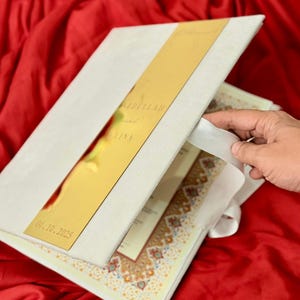 Puede incluir: Una invitación de boda blanca con un acento metálico dorado. La invitación presenta los nombres "Abdullah y Lina" y la fecha "04.10.2025". La invitación está abierta, revelando páginas impresas decorativas.