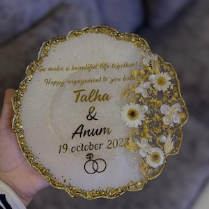 May include: A round, white engagement coaster with gold glitter edging and floral accents. The coaster has the names "Talha & Anum" and the date "19 october 2025" in gold script, along with the text "Go make a beautiful life together! Happy engagement to you both."