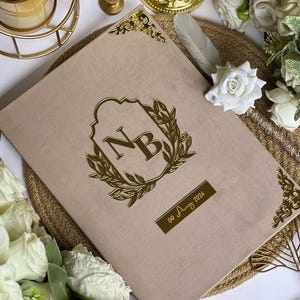 May include: A beige wedding invitation with gold accents. The cover features a gold emblem with the initials "NB" and a laurel wreath. The date "09 Aug 2024" is displayed on a gold rectangular plaque. Decorative gold corner pieces add elegance.