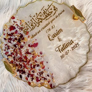 May include: A decorative, round resin tray with gold trim and handles. The tray features gold Arabic calligraphy and the English text "And we created you in pairs (78:8) Qasim & Fatima 30-07-2025". It is decorated with red rose petals and gold flakes.
