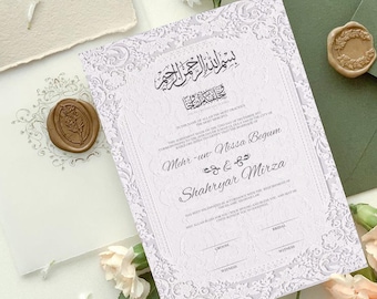 May include: A white wedding invitation with ornate floral detailing and calligraphy. The invitation includes the names Mehr-un Nissa Begum and Shahryar Mirza. A wax seal and flowers are also visible.
