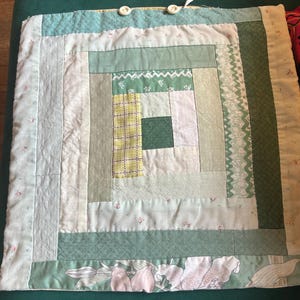 May include: A square, handmade quilt with a log cabin design. The quilt features various shades of green and white fabrics, including floral prints, plaid, and solid colours. The edges are finished with a green floral fabric and the top has two white buttons.