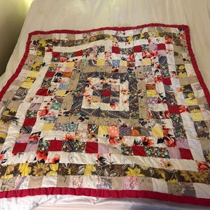 May include: A patchwork quilt with a red border. The quilt is made of various fabric squares in floral and solid patterns, including red, yellow, white, and brown. The quilt is laid out on a white bed.