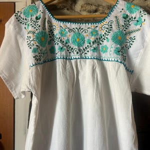 May include: A white short-sleeved blouse featuring a square neckline and intricate embroidery across the chest. The embroidery consists of teal floral patterns with yellow centres, accented by green vines and leaves, all stitched onto the white fabric.