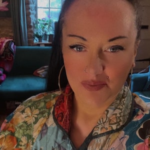 May include: A person wearing a vibrant patchwork jacket with floral and geometric designs. The jacket incorporates pink, blue, green, and red hues. Large gold hoop earrings are worn. A window and a green sofa are in the background.