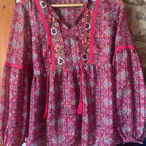 May include: A magenta blouse with long, gathered sleeves and a V-neck. It features a detailed floral embroidery, pink trim, and tassel ties. The patterned fabric hangs on a wooden hanger, showcasing the blouse's design and style.