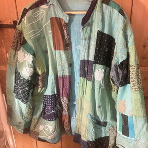 May include: A patchwork jacket in shades of teal, brown, and black, hanging on a wooden hanger. The jacket features a variety of fabric patterns and textures, with a button-up front and two pockets. The overall style is bohemian and unique.