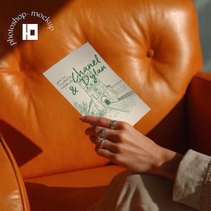 以下が含まれることがあります： 「Chanel & Dylan」と書かれた緑色の文字と建物の絵が描かれたクリーム色の招待状。オレンジ色の革張りの椅子の前に招待状が掲げられています。「you're invited to celebrate」という文字も招待状に書かれています。