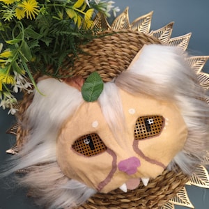 Op de afbeelding: Een handgemaakt leeuwenmasker met een beige gezicht, wit en grijs haar en bruine gaasogen. Het masker heeft een roze neus en mond en witte tanden. Het is geplaatst op een geweven mand met gouden bladaccenten, met gele en groene bloemaccenten.