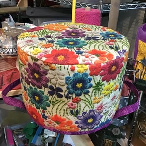 May include: A round, floral-patterned storage container with a purple handle. The container is covered in a vibrant array of embroidered flowers in shades of red, orange, purple, blue, and yellow. The background is a cream color.