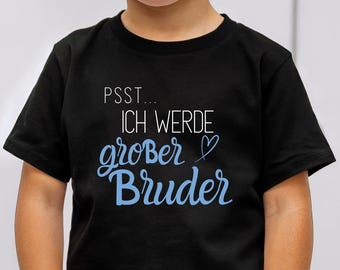 May include: A young child wearing a black t-shirt with the text "PSST... ICH WERDE großer Bruder" in blue script. The child is wearing blue jeans and has their hands in their pockets. The shirt design features a heart symbol.
