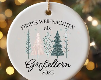 May include: A white ceramic Christmas ornament with a gold string for hanging. The ornament features the text "Erstes Weihnachten als Großeltern 2025" and illustrations of Christmas trees in teal, pink, and green.