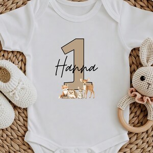 May include: White baby onesie with a tan number 1 and the name "Hanna" in black script. Below the text are illustrations of woodland animals. A pair of white baby shoes and a crochet bunny rattle are also in the image.