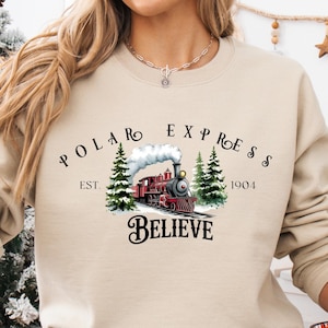 May include: Beige sweatshirt featuring a graphic of a red train with the words "POLAR EXPRESS" and "BELIEVE". The design includes two green pine trees and the year "1904". A festive holiday-themed apparel item.