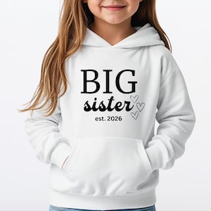 Puede incluir: Una sudadera con capucha blanca con el texto negro "BIG sister est. 2026". La sudadera tiene un bolsillo delantero y la lleva una niña pequeña con gafas de sol blancas.