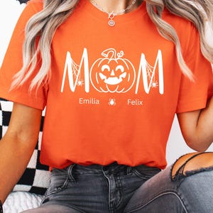 Puede incluir: Camiseta naranja con un diseño blanco con temática de Halloween. El diseño incluye la palabra "MOM" con una calabaza y telarañas, y los nombres "Emilia" y "Felix" debajo. La camiseta es de cuello redondo y manga corta.