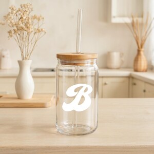 May include: A clear glass tumbler with a wooden lid and a glass straw. The tumbler features a white letter "B" on the front. The tumbler is sitting on a light-colored wooden surface.