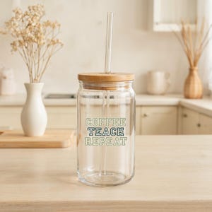 May include: A clear glass tumbler with a wooden lid and a glass straw. The tumbler has the words "COFFEE TEACH REPEAT" in green and white lettering. The tumbler is sitting on a light-colored wooden surface.