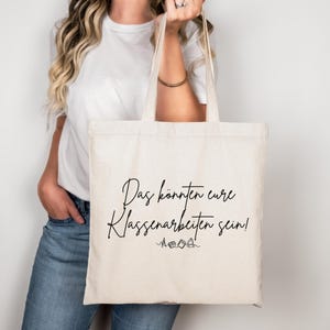 Può includere: Una borsa tote color crema con scritta nera che recita "Das könnten eure Klassenarbeiten sein!". La borsa è tenuta da una persona che indossa una camicia bianca e jeans blu. La borsa ha un design semplice.