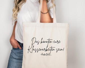 May include: A cream-colored tote bag with black script that reads "Das könnten eure Klassenarbeiten sein!" The bag is held by a person wearing a white shirt and blue jeans. The bag has a simple design.