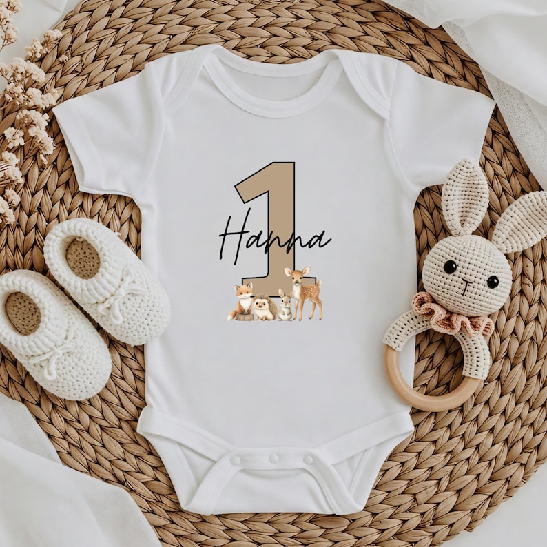 May include: White baby onesie with a tan number 1 and the name "Hanna" in black script. Below the text are illustrations of woodland animals. A pair of white baby shoes and a crochet bunny rattle are also in the image.