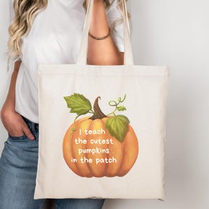 May include: Cream-colored tote bag featuring a large orange pumpkin illustration with green leaves and the text "I teach the cutest pumpkins in the patch." The bag has two handles.