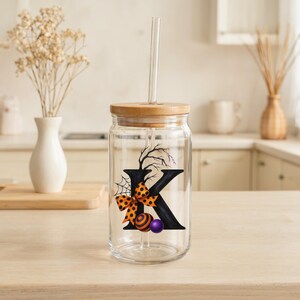 May include: A clear glass tumbler with a bamboo lid and a glass straw. The tumbler features a black letter "K" decorated with a spiderweb, a bow with orange and black polka dots, and two decorative balls. The tumbler is on a light-colored wooden surface.