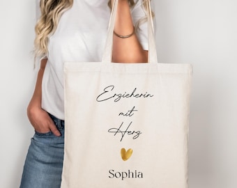 May include: Cream-colored tote bag with black script text that reads "Erzieherin mit Herz" and the name "Sophia" below a gold heart. The bag has two handles and is held by a person wearing a white shirt and blue jeans.