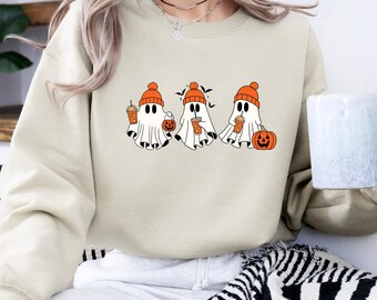 May include: A beige sweatshirt featuring a Halloween-themed graphic. The graphic depicts three cartoon ghosts wearing orange beanies, holding coffee cups, and accompanied by pumpkins and bats. The sweatshirt is a neutral color.