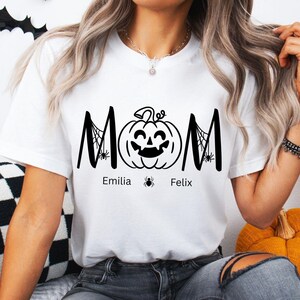 Puede incluir: Camiseta blanca con un diseño negro con temática de Halloween. La palabra "MOM" se muestra con una calabaza en el centro. Los nombres "Emilia" y "Felix" están impresos debajo de la calabaza, con gráficos de arañas.